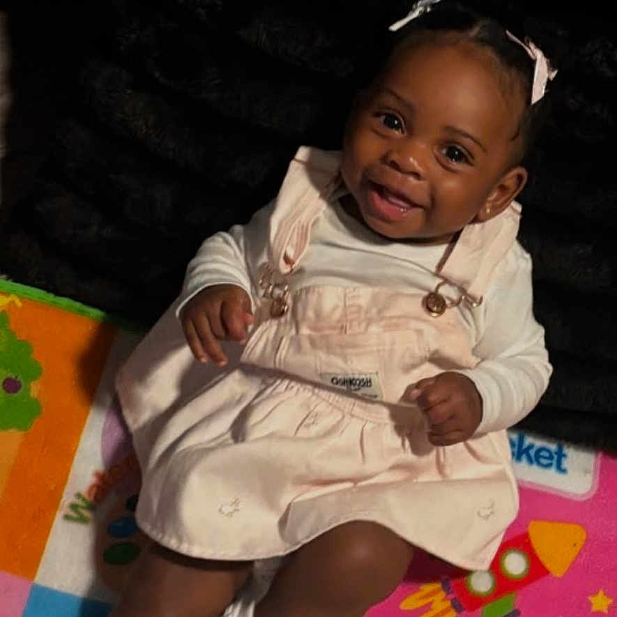 Janiyah Rhodes is registered to the contest to win money with this photo: baby, child, infant, smiling, portrait, sitting, indoor, play_mat, colorful, dress, overalls, sneakers, hair_bows, dark_skin, happy, playroom, toy_mat, closeup, feet, casual