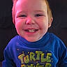 child, toddler, smiling, sweatshirt, blue_clothing, text_on_clothing, indoor, portrait, happy, face, person, casual_wear, cute, young_child, seated, home, lighting, colorful, expression, sitting