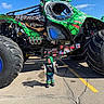 Edward joined the competition — help win amazing prizes! child, monster_truck, vehicle, tire, outdoor, parking_lot, blue_sky, green, black, clothing, jeans, boots, hoodie, sunny, shadow, concrete, large_vehicle, small_child, funny_size_contrast, daytime