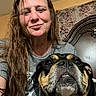 woman, dog, indoor, portrait, long_hair, gray_shirt, brown_hair, headboard, curtain, face, pet, closeup, smile, expression, furniture, casual, home, resting, companion, candid