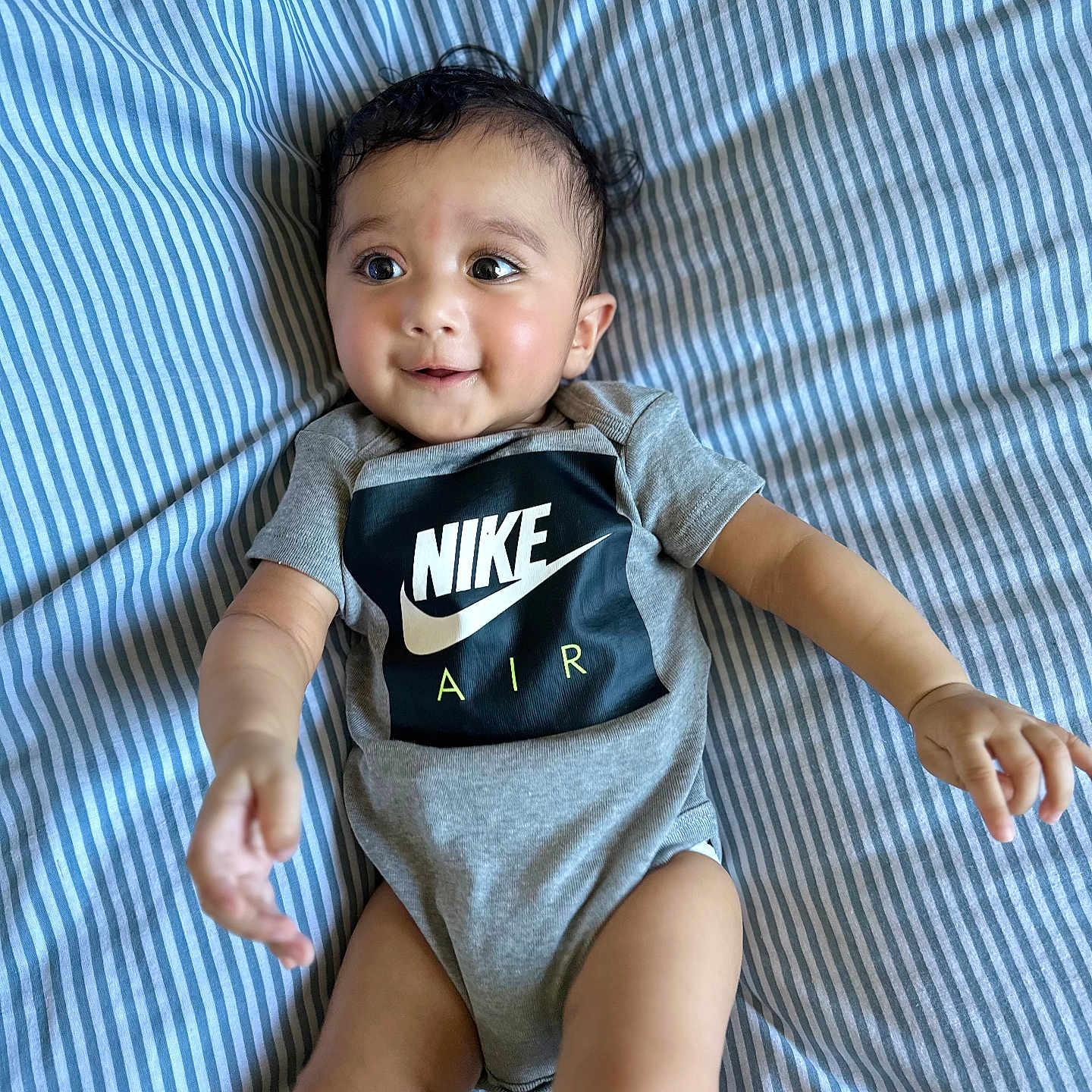 Risha joined the competition — help win amazing prizes! arms_outstretched, baby, blue_and_white, child, clothing, curious, cute, face, happy, indoor, infant, lying_down, nike_air, onesie, person, portrait, skin, smiling, striped_bed_sheet, young_child