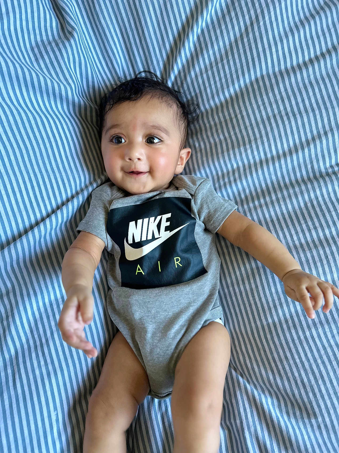 Risha joined the competition — help win amazing prizes! baby, infant, child, onesie, nike_air, striped_bed_sheet, blue_and_white, smiling, happy, curious, lying_down, indoor, cute, young_child, arms_outstretched, clothing, portrait, face, person, skin