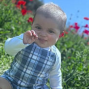 Marcus is registered to the contest to win money with this photo: baby, child, plaid_romper, grass, flowers, red_flowers, outdoor, sunny, greenery, blue_sky, cute, playful, sitting, nature, infant, portrait, smiling, happy, one_person, young_child