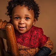Kiyeir is registered to the contest to win money with this photo: toddler, child, smiling, curly_hair, knit_sweater, red_sweater, orange_sweater, black_sweater, wooden_chair, portrait, happy, cute, indoors, warm_lighting, close_up, person, baby, face, eyes, expression