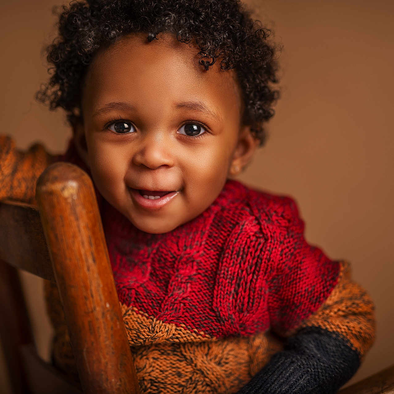 Kiyeir is registered to the contest to win money with this photo: baby, black_sweater, child, close_up, curly_hair, cute, expression, eyes, face, happy, indoors, knit_sweater, orange_sweater, person, portrait, red_sweater, smiling, toddler, warm_lighting, wooden_chair