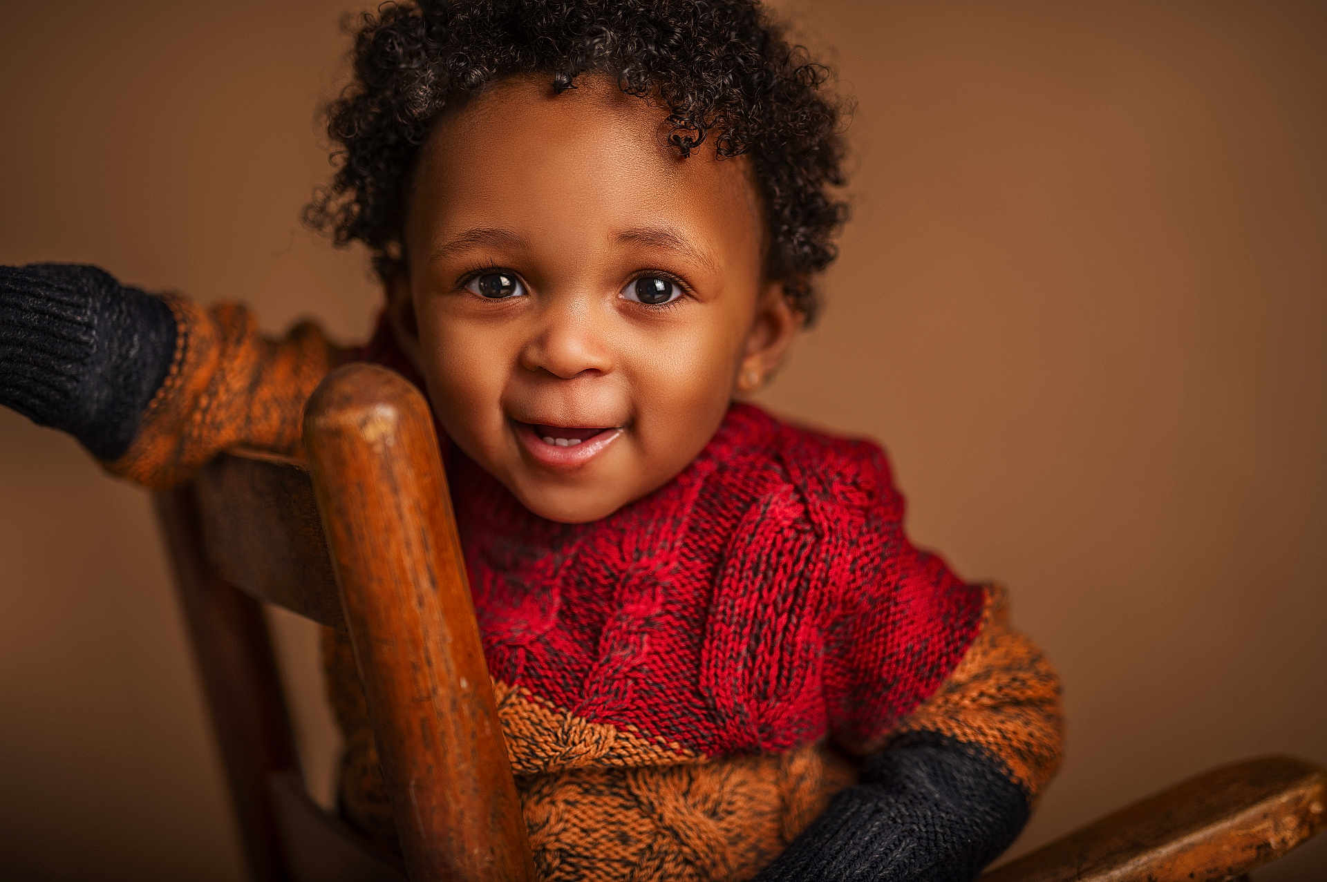Kiyeir is registered to the contest to win money with this photo: toddler, child, smiling, curly_hair, knit_sweater, red_sweater, orange_sweater, black_sweater, wooden_chair, portrait, happy, cute, indoors, warm_lighting, close_up, person, baby, face, eyes, expression