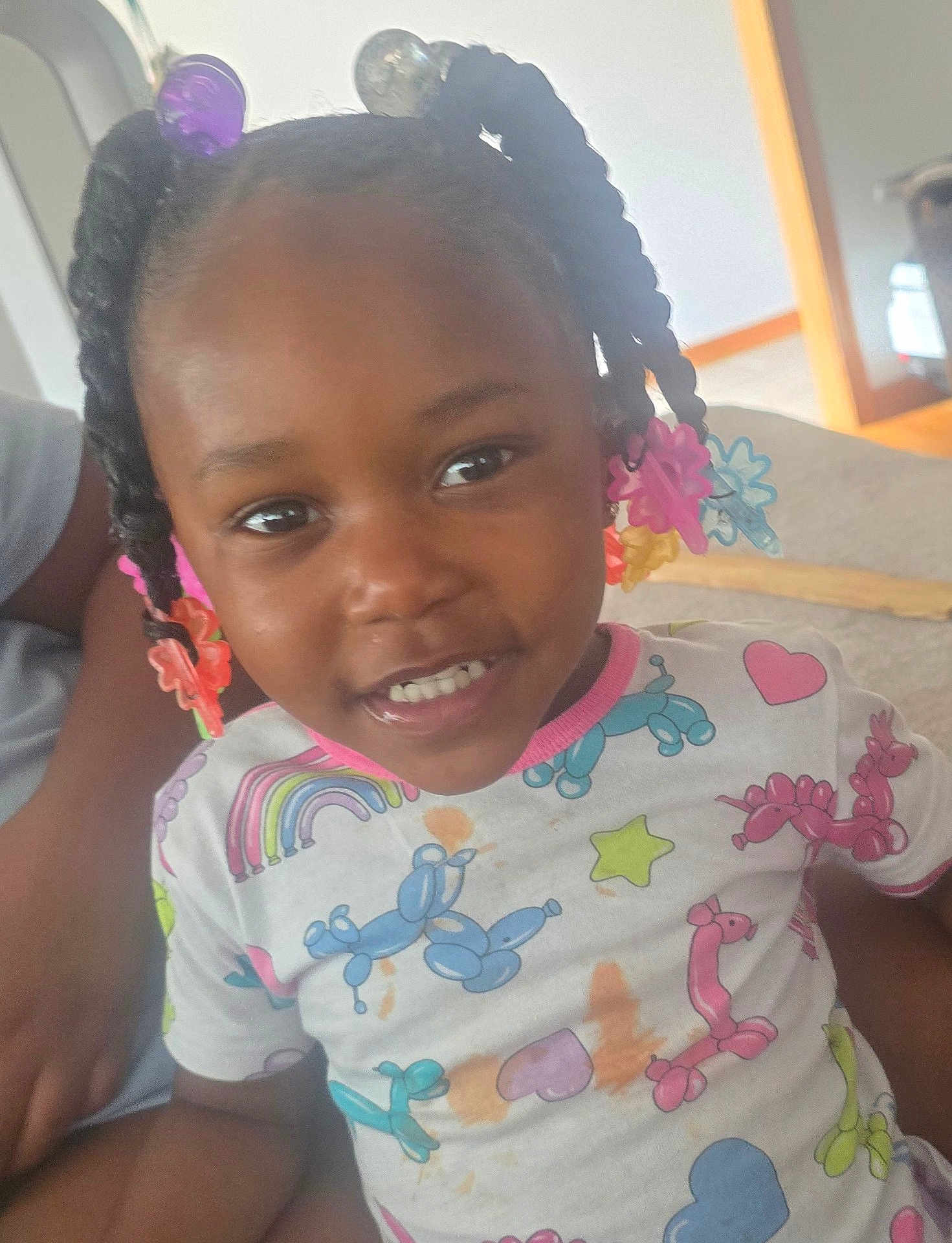 Yani is registered to the contest to win money with this photo: child, smiling, hair_accessories, braids, balloon_animal_print, shirt, indoor, person, cute, happy, seated, colorful, toddler, portrait, casual, playful, young_child, face, clothing, home