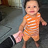 baby, baby_gate, barefoot, bright_colors, carpet, child, couch, curly_hair, hand_holding, home, indoor, living_room, orange_clothing, person, portrait, smiling, standing, striped_shirt, support, toddler