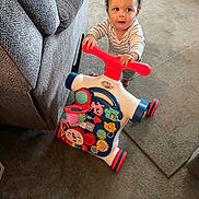 Braxton is registered to the contest to win money with this photo: balloons, barefoot, carpet, child, colorful_toy, couch, curly_hair, floor, furniture, home, indoor, living_room, orange_cushion, person, play, smiling, striped_clothing, toddler, toy_walker, young_child