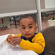 Jaydën participe au concours pour gagner de l'argent avec cette photo : child, toddler, boy, food, bread, table, indoor, sitting, yellow_sweater, face, eyes, hands, snack, crumbs, curious, expression, chair, window, plant, casual