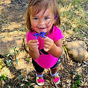 Sofia is registered to the contest to win money with this photo: child, girl, flower, pink_shirt, sandals, outdoor, nature, smiling, happy, greenery, rocks, leaves, sunlight, shorts, playful, portrait, young_child, casual_clothing, holding, summer