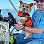 dog, corgi, life_jacket, man, boat, steering_wheel, water, lake, sky, sunglasses, cap, smile, casual_clothing, watch, jeans, outdoor, pet, holding, seated, happy