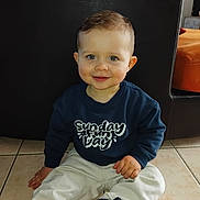 Tiago a rejoint le concours — aidez-le/la à gagner de superbes lots ! child, toddler, baby, face, blue_eyes, smile, sitting, tiled_floor, navy_sweatshirt, white_pants, navy_shoes, toy, leather_chair, indoor, portrait, short_hair, rosy_cheeks, playful, apparel_text, ear