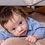 baby, infant, blue_eyes, cushion, soft_rug, blue_shirt, indoor, curious, lying_down, face, hand, hair, pillow, floor, furniture_leg, skin, child, closeup, portrait, home