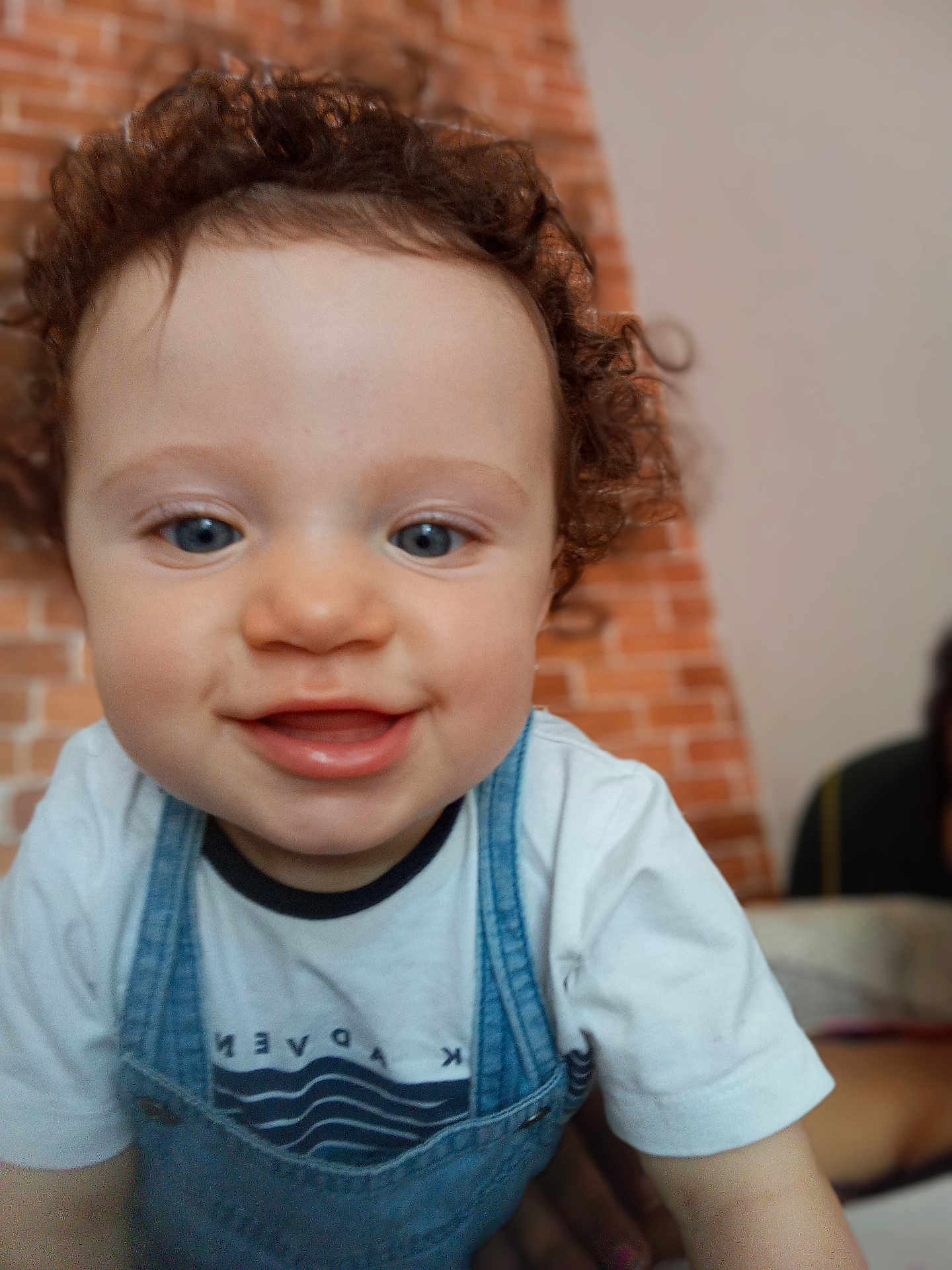 Gabriel a rejoint le concours — aidez-le/la à gagner de superbes lots ! toddler, child, curly_hair, smiling, blue_eyes, denim_overalls, white_tshirt, close_up, indoor, brick_wall, happy, person, cute, face, portrait, baby, young_child, curiosity, soft_focus, background_blur