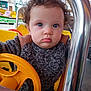 Gabriel a rejoint le concours — aidez-le/la à gagner de superbes lots ! toddler, child, curly_hair, blue_eyes, yellow_steering_wheel, amusement_ride, sweater, metal_bar, outdoor, face, portrait, serious_expression, seat, playground, person, clothing, hands, vehicle, daylight, cute