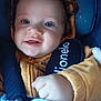 baby, child, face, smile, blue_eyes, curly_hair, hoodie, mustard_color, car_seat, seatbelt, blanket, hand, close_up, infant, cute, portrait, young_child, warm_clothing, happy, indoor