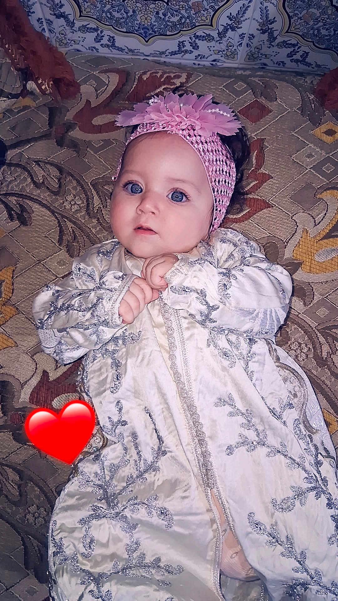 Sarah participe au concours pour gagner de l'argent avec cette photo : baby, child, blue_eyes, pink_headband, flower_headband, white_outfit, embroidered_clothing, carpet, patterned_carpet, indoor, portrait, cute, infant, lying_down, hand_position, red_heart_emoji, decorative, closeup, person, young
