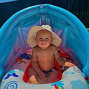 Josiah joined the competition — help win amazing prizes! baby, child, inflatable_pool, pool_float, hat, sun_hat, water, summer, outdoor, sunlight, blue_sky, fence, sitting, cute, smiling, playful, colorful, fun, vacation, warm_weather