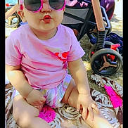 Taqdeer is registered to the contest to win money with this photo: toddler, child, sunglasses, pink_shirt, shorts, blanket, stroller, outdoor, sunlight, footwear, sandals, grass, playtime, summer, cute, pucker, baby, sitting, funny_face, accessory