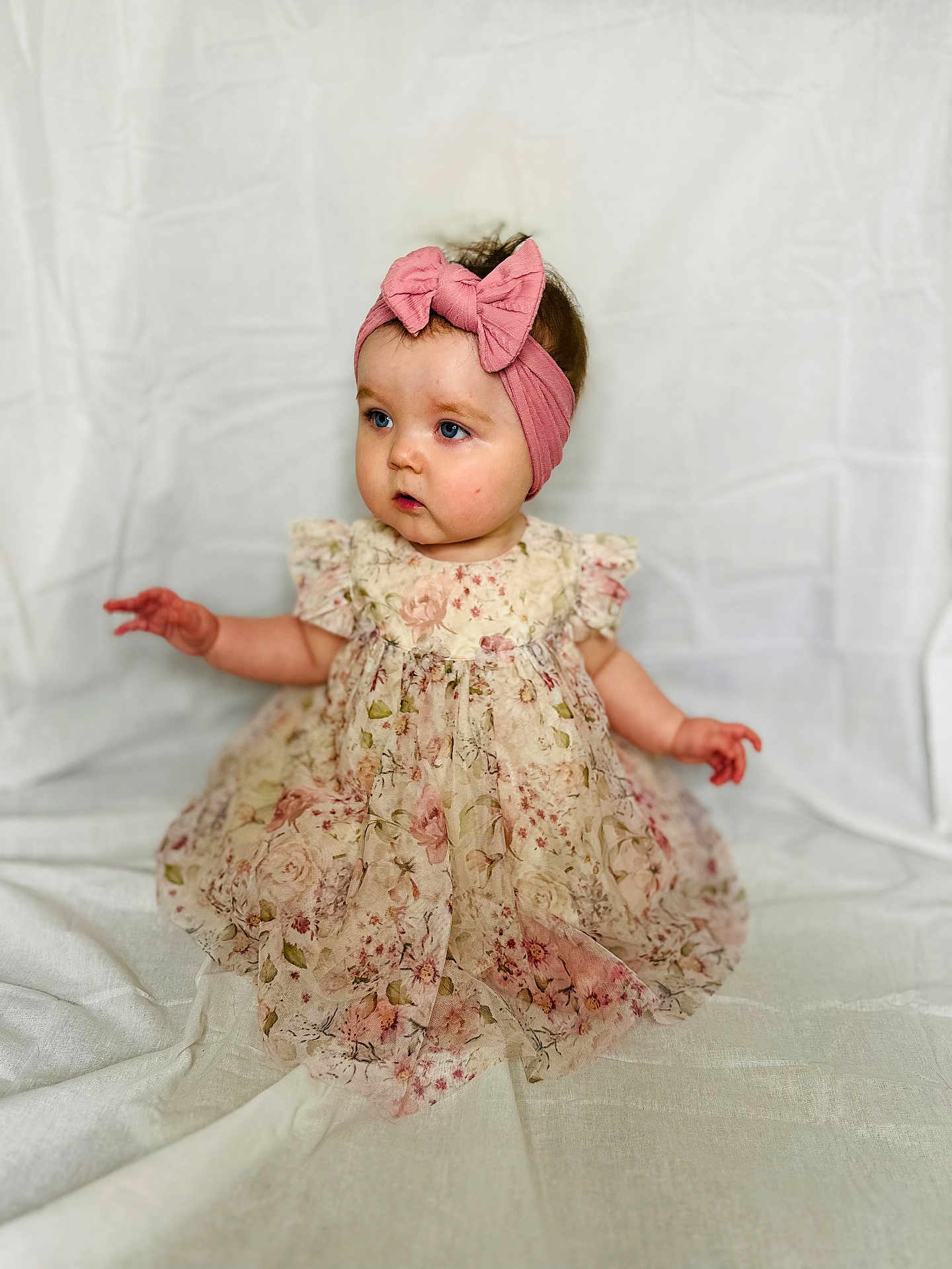Nora-Irene is registered to the contest to win money with this photo: apparel, baby, blue_eyes, bow, child, curious, cute, dress, fabric, floral, girl, headband, indoors, infant, pattern, pink, portrait, sitting, soft_background, young