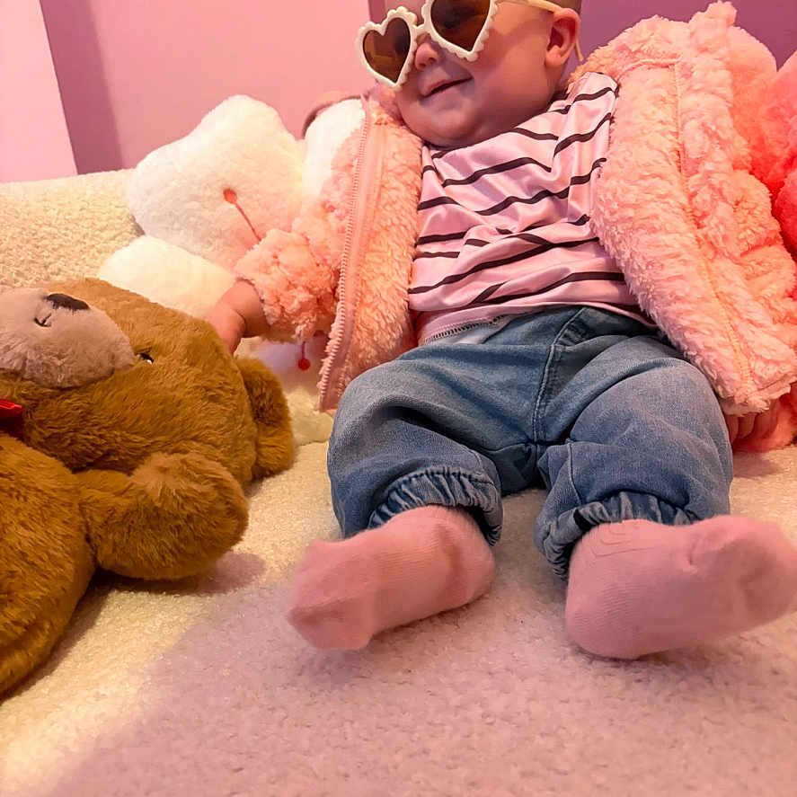 Lyra is registered to the contest to win money with this photo: baby, child, heart_shaped_sunglasses, pink_jacket, striped_shirt, blue_jeans, pink_socks, teddy_bear, plush_toy, cozy, indoor, cute, smiling, seated, soft_texture, childrens_room, fashion, relaxed, portrait, infant