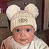 Lyra is registered to the contest to win money with this photo: baby, child, hat, pom_poms, blue_eyes, indoor, cute, knitwear, infant, face, portrait, sweater, furniture, cabinet, floor, warm_clothing, expression, adorable, headwear, room