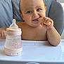 baby, high_chair, bottle, food, snack, child, face, hand, blue_eyes, table, crumbs, smile, indoor, cute, feeding, happy, person, closeup, seat, toddlers