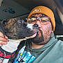 man, dog, brindle_dog, beanie, glasses, hoodie, naruto, selfie, close_up, car_interior, facial_hair, tape, hand, collar, pet, nuzzling, affection, casual_clothing, indoors, portrait