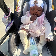 Nyziah is registered to the contest to win money with this photo: baby, infant, stroller, car_seat, headband, bow, bottle, socks, tutu, dress, sitting, hand_in_mouth, toy, plush_toy, safety_label, sunlight, outdoor, pavement, closeup, child