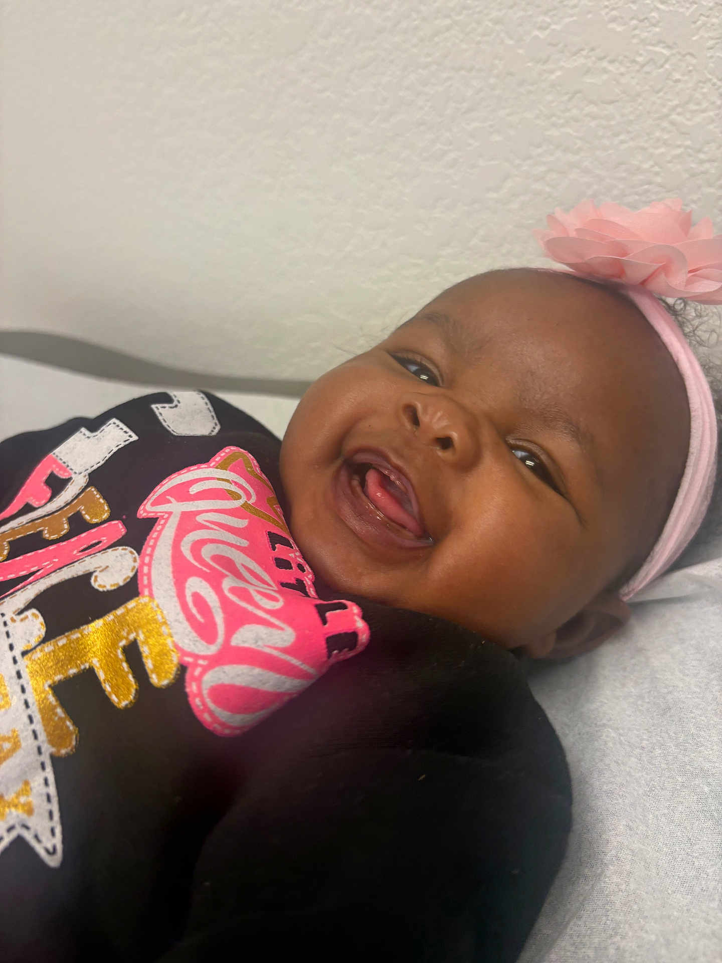 Nyziah joined the competition — help win amazing prizes! baby, infant, smiling, laughing, headband, pink_flower, closeup, portrait, dark_skin, tongue, teeth, onesie, colorful_print, wall, sheet, face, cheeks, eyes, happy, cute