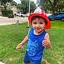 child, toddler, boy, fireman_hat, blue_tank_top, shorts, sandwich, grass, sidewalk, suburban, trees, cars, smiling, outdoor, daytime, playful, street, clothing, person, happy