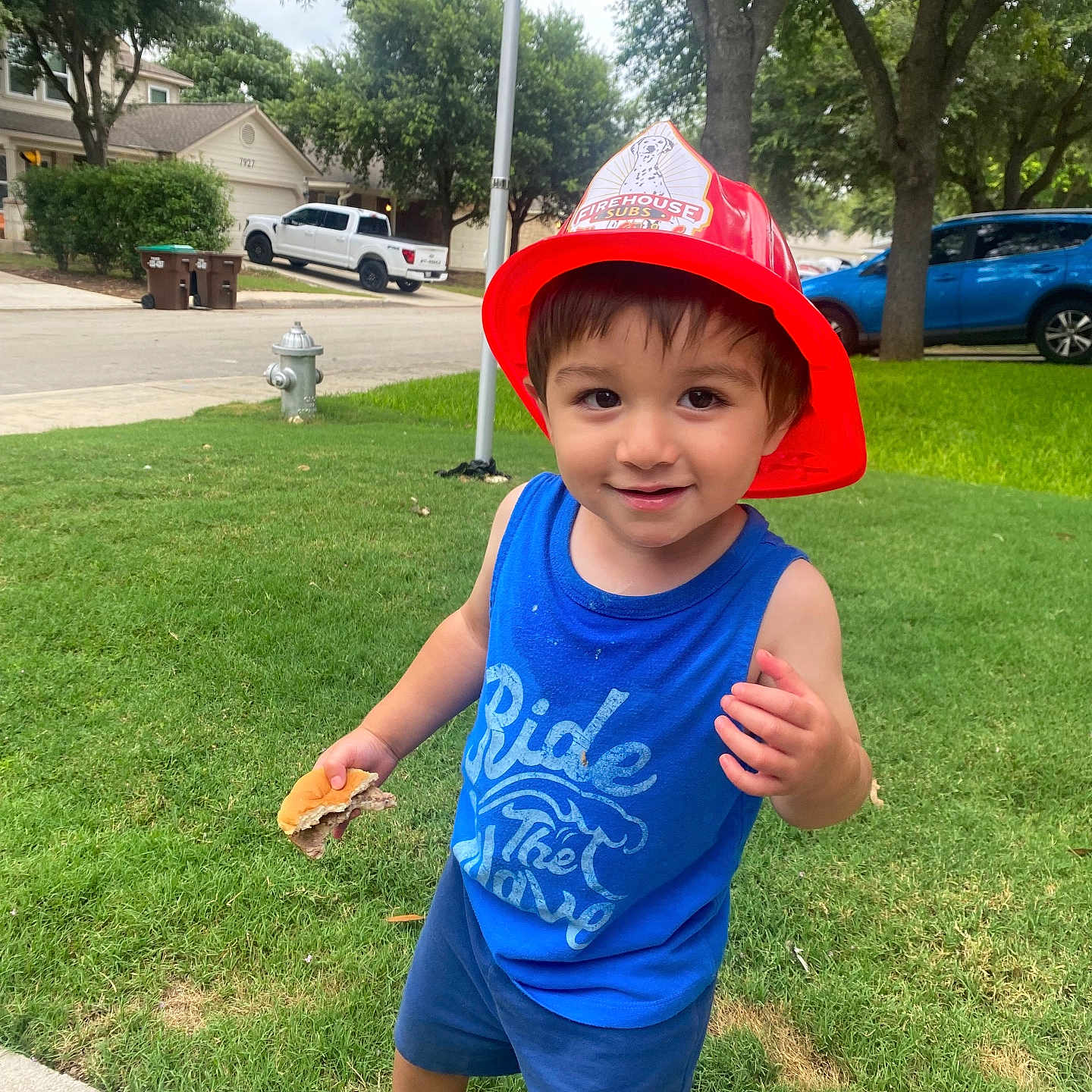 Ian Elijah is registered to the contest to win money with this photo: blue_tank_top, boy, cars, child, clothing, daytime, fireman_hat, grass, happy, outdoor, person, playful, sandwich, shorts, sidewalk, smiling, street, suburban, toddler, trees