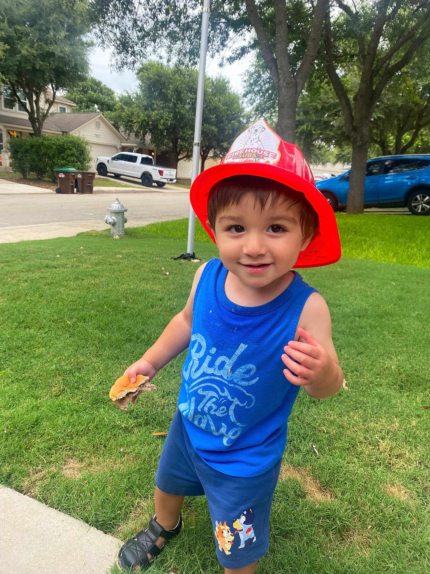 Ian Elijah is registered to the contest to win money with this photo: child, toddler, boy, fireman_hat, blue_tank_top, shorts, sandwich, grass, sidewalk, suburban, trees, cars, smiling, outdoor, daytime, playful, street, clothing, person, happy