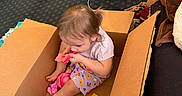 Kristie joined the competition — help win amazing prizes! child, toddler, cardboard_box, toy, play_food, indoor, carpet, clothing, person, sitting, brown_box, pink, blue_toy, floor, hair, foot, living_room, furniture, couch, playtime