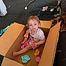 child, cardboard_box, toys, indoor, carpet, baby_swing, playtime, curious, casual_clothing, footwear, plastic_toys, living_room, messy_room, young_child, sitting, barefoot, playful, household_items, candid, childhood