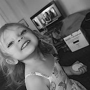 Kristie is registered to the contest to win money with this photo: black_and_white, child, cute, dress, face, furniture, girl, hair, happy, home, indoor, living_room, person, playful, portrait, smiling, snack, television, toddler, young