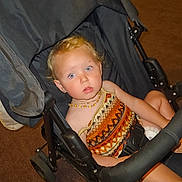 Mayana participe au concours pour gagner de l'argent avec cette photo : child, toddler, stroller, indoor, carpet, blonde_hair, blue_eyes, necklace, patterned_clothing, seated, black_stroller, curly_hair, baby, person, expression, looking_at_camera, casual, infant, cute, young_child