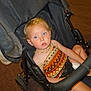 child, toddler, stroller, indoor, carpet, blonde_hair, blue_eyes, necklace, patterned_clothing, seated, black_stroller, curly_hair, baby, person, expression, looking_at_camera, casual, infant, cute, young_child