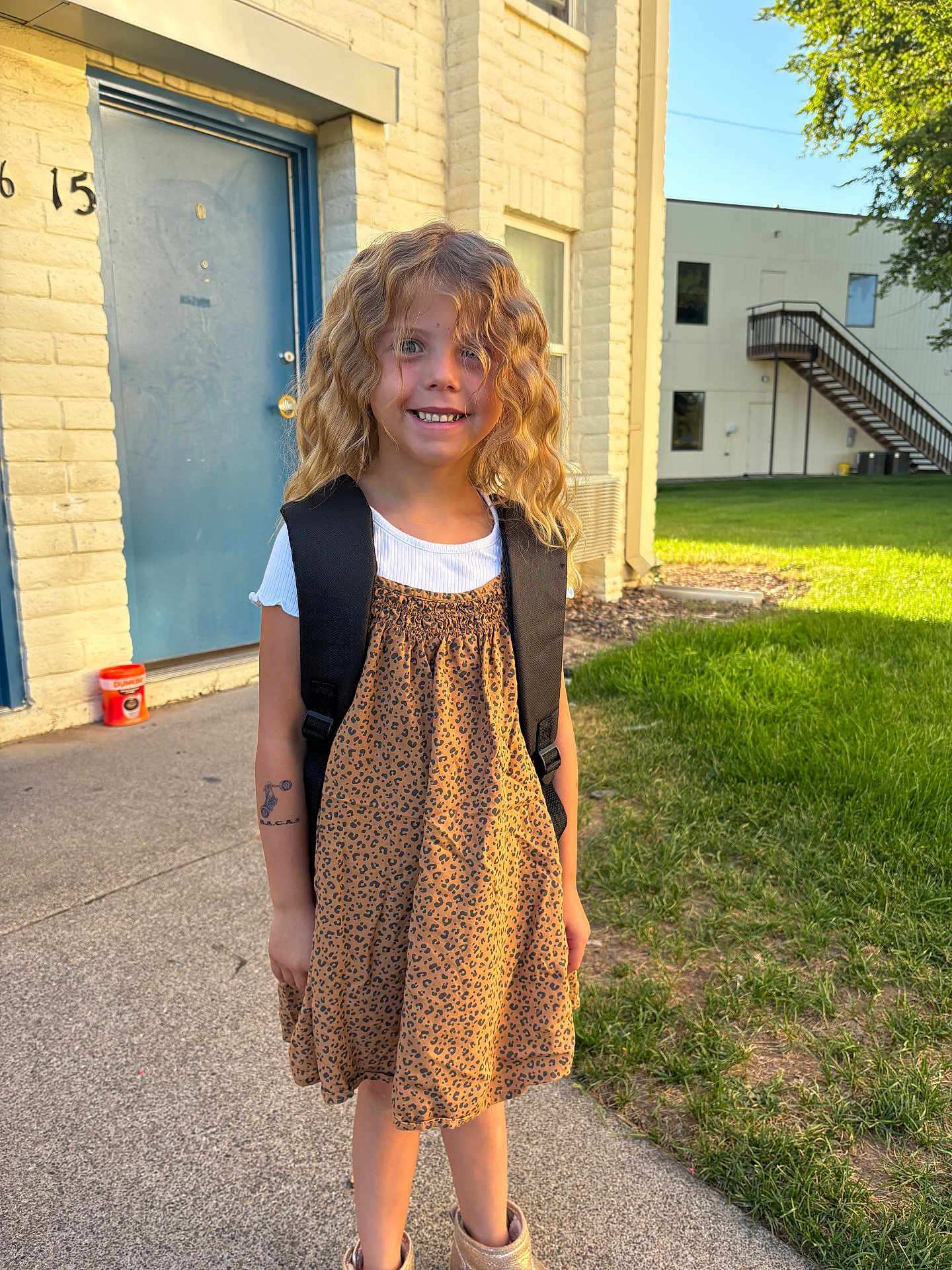 Serenity joined the competition — help win amazing prizes! child, girl, smiling, curly_hair, blonde_hair, leopard_print, dress, backpack, white_shirt, outdoor, sidewalk, grass, building, door, window, sunlight, happy, tattoo, footwear, daylight