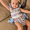 baby, beige, blue_checkered_bow, child, couch, cute, floral_pattern, flower_headband, footwear, girl, happy, headband, indoor, person, portrait, romper, sitting, skin, smiling, young