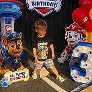Eli is registered to the contest to win money with this photo: child, boy, birthday, party, paw_patrol, balloons, cartoon_characters, birthday_sign, number_three, costume, smiling, standing, outdoor, colorful, decorations, toy_dog, firefighter_dog, police_dog, pavement, portrait