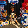 child, boy, birthday, party, paw_patrol, balloons, cartoon_characters, birthday_sign, number_three, costume, smiling, standing, outdoor, colorful, decorations, toy_dog, firefighter_dog, police_dog, pavement, portrait