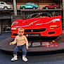 automobile, baby, car, child, clothing, curious, display, exhibit, ferrari, floor, head, indoor, jeans, museum, person, red_car, sneakers, toddler, vehicle, vintage_car