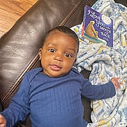 Kaylx joined the competition — help win amazing prizes! baby, child, blue_onesie, brown_leather_couch, blanket, book, floor, wood_floor, smiling, face, person, indoor, infant, cute, portrait, lying_down, cozy, reading, expression, home