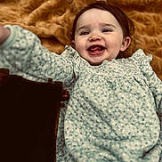 Everleigh is registered to the contest to win money with this photo: baby, blanket, child, clothing, comfort, cozy, cute, face, floral_dress, happy, indoors, infant, lying_down, person, portrait, smiling, soft_texture, tongue_out, warm_colors, young_child