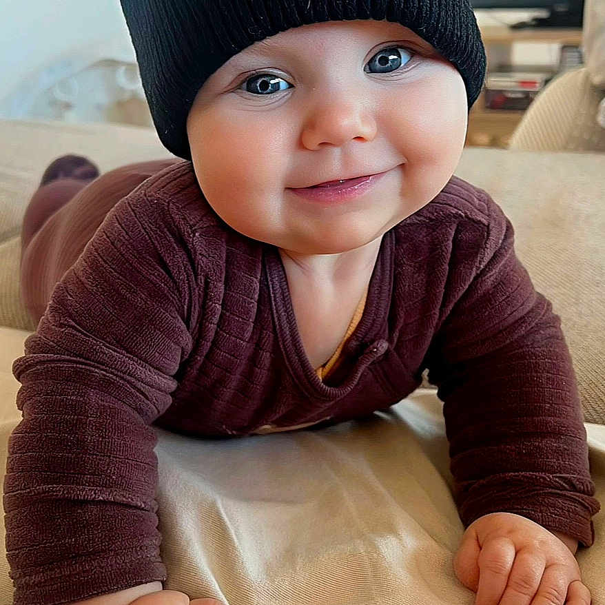 Phœbe participe au concours pour gagner de l'argent avec cette photo : baby, black_hat, blanket, blue_eyes, brown_clothing, child, cozy, cute, face, hand, happy, hat, indoors, infant, pacifier, person, pom_pom, portrait, smiling, tummy_time