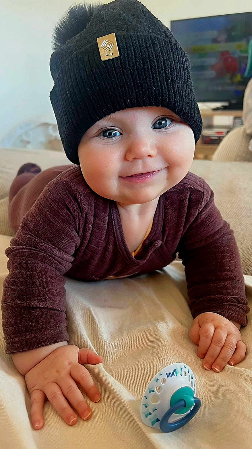 Phœbe participe au concours pour gagner de l'argent avec cette photo : baby, black_hat, blanket, blue_eyes, brown_clothing, child, cozy, cute, face, hand, happy, hat, indoors, infant, pacifier, person, pom_pom, portrait, smiling, tummy_time