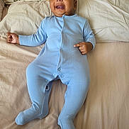 Teeian is registered to the contest to win money with this photo: baby, infant, blue_onesie, bed, pillow, floral_pattern, smiling, pointing, happy, lying_down, indoors, child, person, cute, face, skin, head, sleepwear, comfort, cozy
