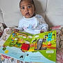 baby, child, book, reading, bed, onesie, floral_pattern, indoor, curious, wide_eyes, colorful, illustration, storybook, hand, face, portrait, seated, young_child, expression, cozy